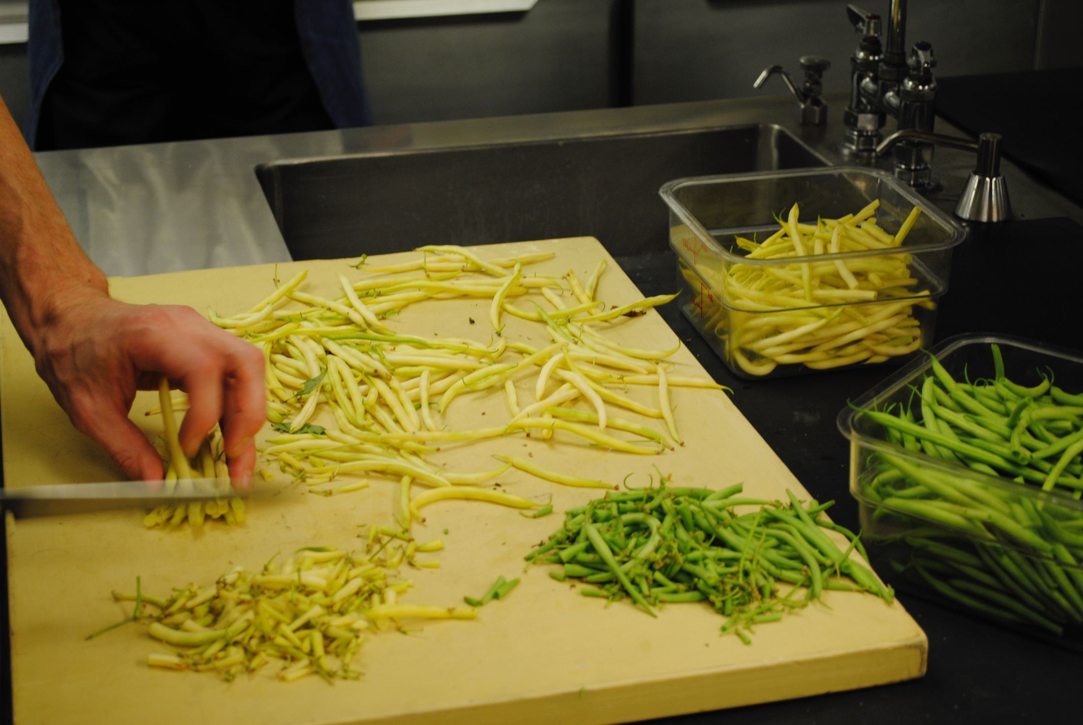 Hands prepping yellow and green beans on a cutting board in a professional kitchen
