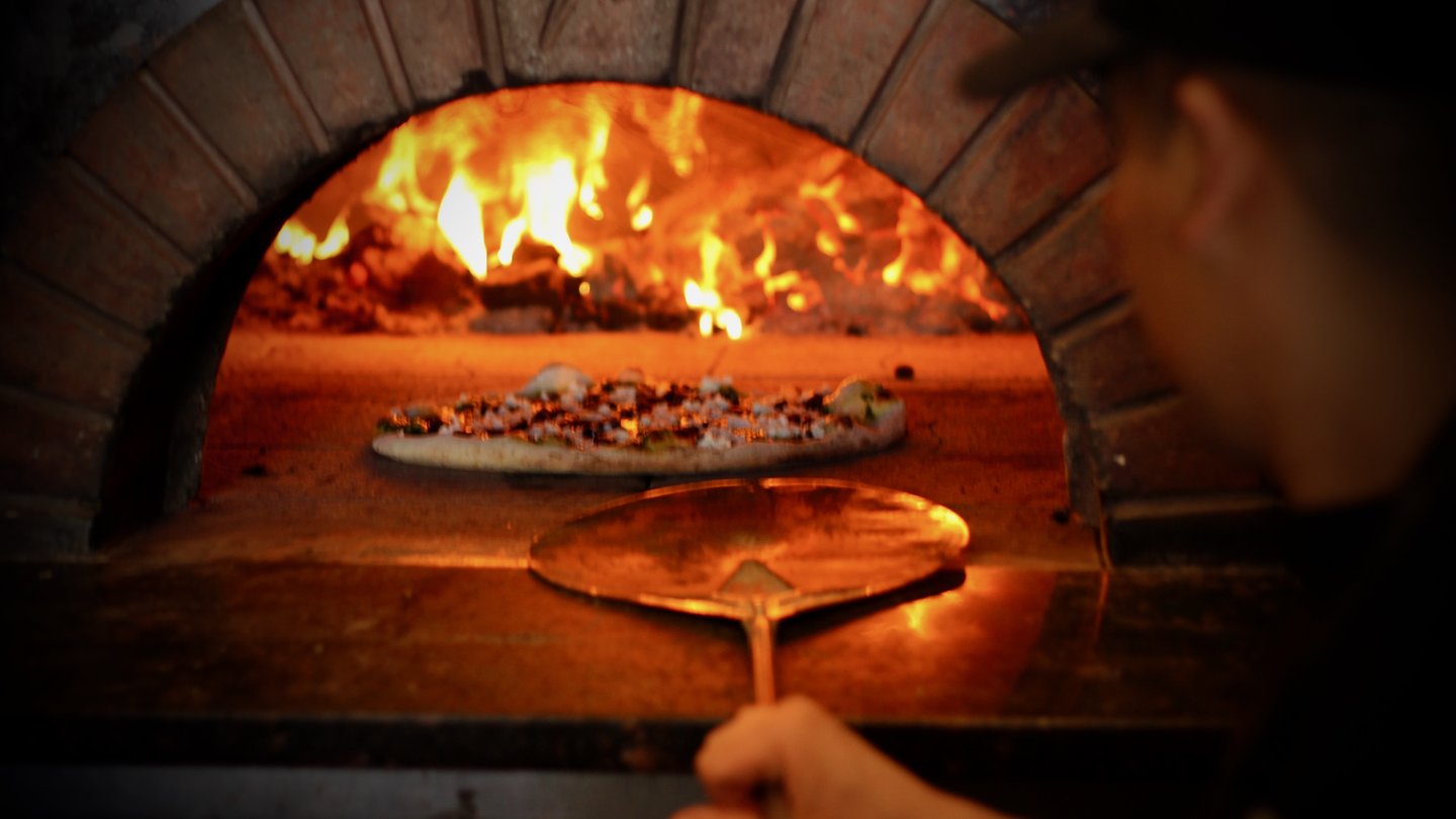 A cook works a pizza peel at a wood-fired oven