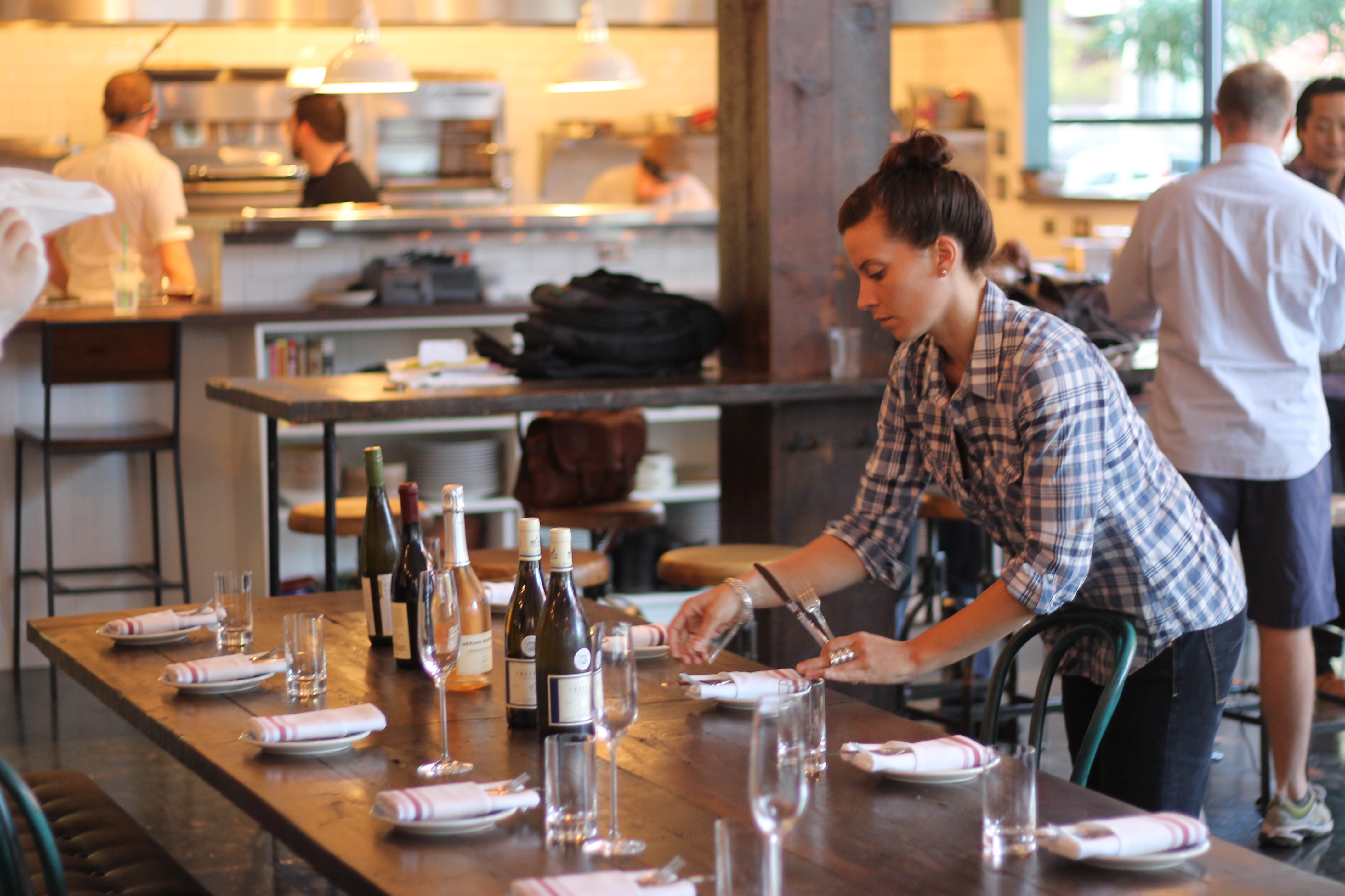 A server setting a long communal table for service, glassware and napkins laid out, open kitchen behind